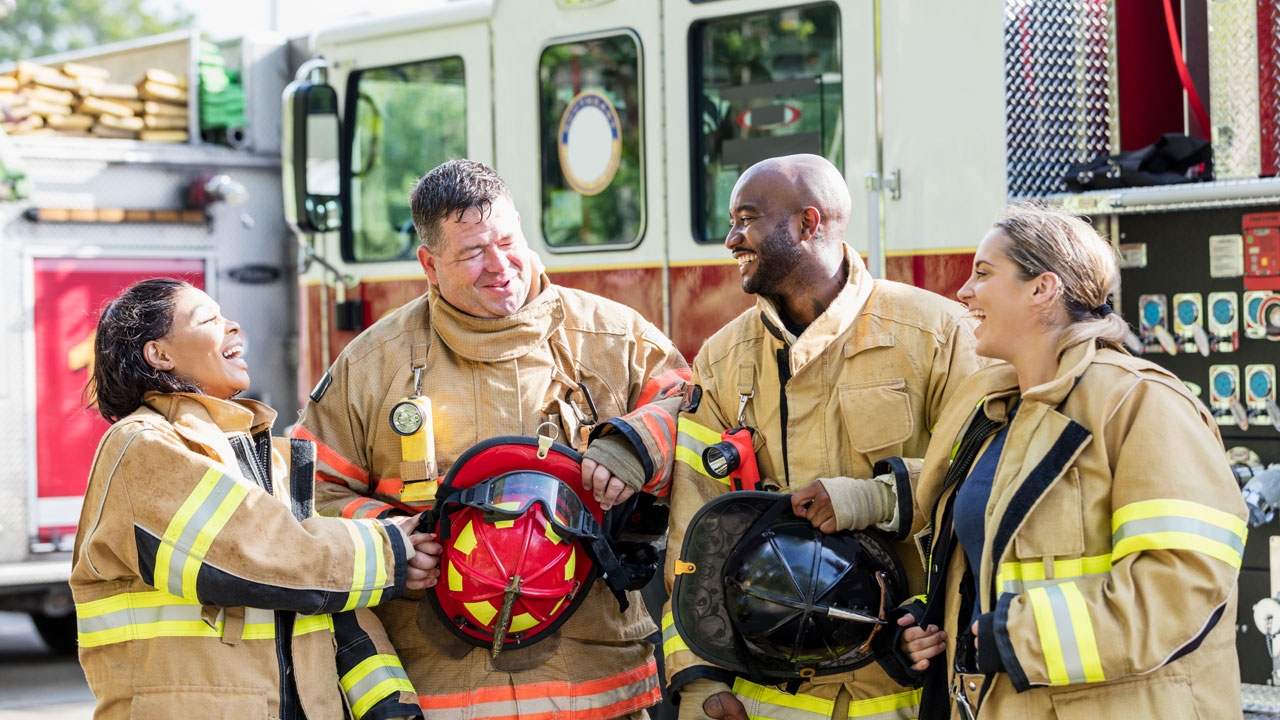 A group of firefighters laughing together with a firetruck in the background